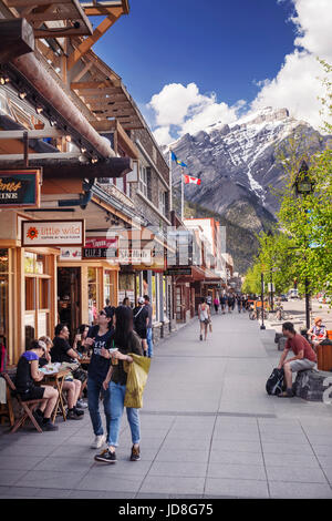 Scenario di strada di persone, negozi e ristoranti su Banff Avenue, nel centro cittadino di Banff in Alberta Rockies con Montagne Rocciose in background. Alberta, Foto Stock