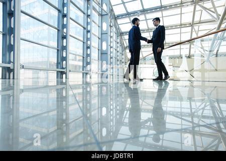 Ampio angolo ritratto di due business contemporaneo uomini stringono le mani nella hall del futuristico edificio di vetro Foto Stock