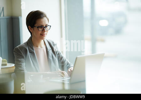 Ritratto di sorridente imprenditrice moderno utilizzando laptop in corrispondenza della finestra tabella in cafe, godendo della pausa caffè Foto Stock