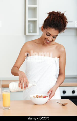 Foto di sorridere giovane donna africana vestita di asciugamano in piedi a casa e guardando in disparte mentre mangiando fragola con muesli. Foto Stock