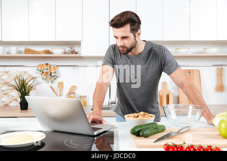 L'uomo alla ricerca di una ricetta su laptop in cucina a casa Foto Stock