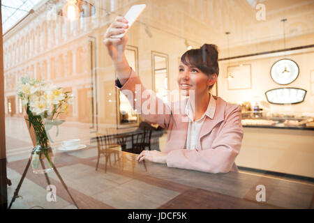 Sorridente giovane donna fare una foto selfie mentre è seduto al tavolo nella caffetteria in ambienti interni Foto Stock