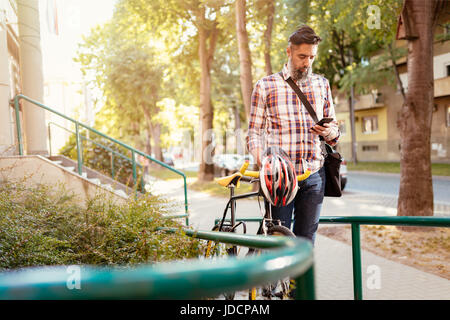 Casual uomo d affari di andare al lavoro in bicicletta. Egli sta spingendo la bicicletta e invio di un messaggio di testo. Foto Stock