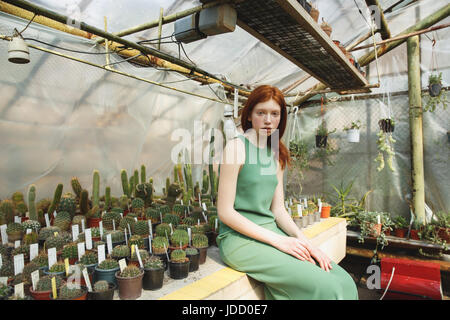 Redhead ragazza in abito verde seduto sul ripiano con cactus e cercando seriamente della fotocamera Foto Stock