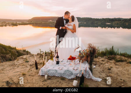 Il kissing sposi sullo sfondo del fiume durante il tramonto dietro il set da pic-nic Foto Stock