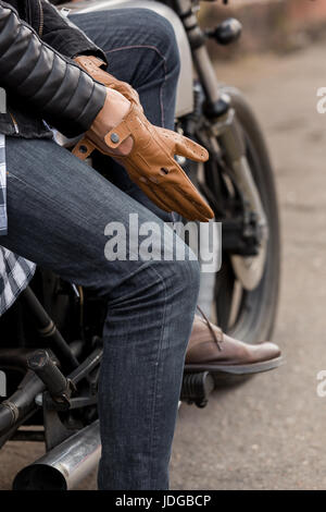 Close-up di un bel rider biker man mano mette su un guanto e sedersi sul classico stile Café Racer motocicletta. Moto custom made in vintage garage. Brut Foto Stock