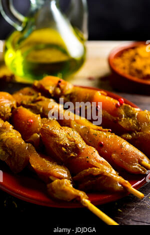 Primo piano di alcune materie Pollo speziato spiedini di carne in un piatto di terracotta, su una tavola in legno rustico, accanto a una ampolla di vetro con olio di oliva e una ciotola wit Foto Stock