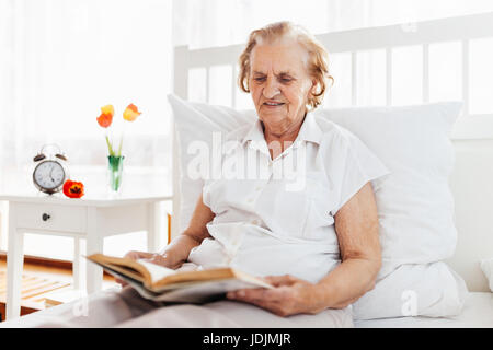 Donna anziana seduti comodamente nel letto leggendo il suo libro preferito a casa Foto Stock