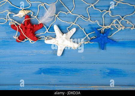 red white and blue starfish in nautical netting with seashell and feather on rustic blue wood Foto Stock