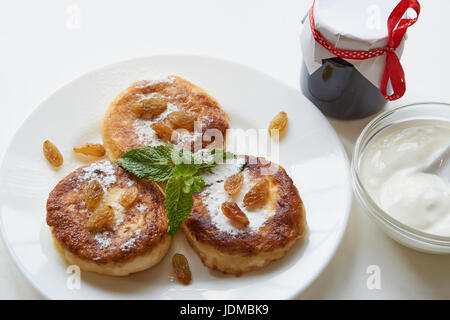 Tre le frittelle su una piastra bianca sul tavolo bianco con la fustigazione di panna acida e un po' di un vasetto di marmellata. ricoperta di zucchero in polvere, menta, uvetta. basso angolo sho Foto Stock