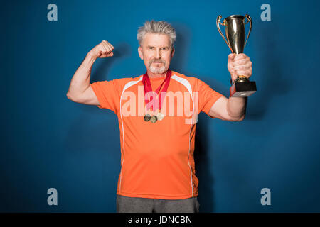 Muscoloso uomo anziano holding trophy e mostrando bicipite isolato su blu Foto Stock