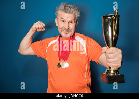 Muscoloso uomo anziano holding trophy e mostrando bicipite isolato su blu Foto Stock