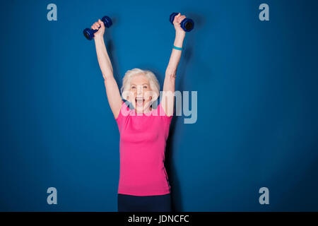 Emozionato senior donna fitness training con manubri isolato su blu Foto Stock