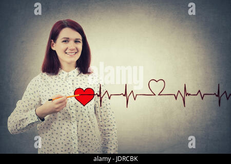 Giovane ragazza sorridente e tenendo una matita per torace, il disegno di un cuore rosso con la sua cardiogram heartbeat. La carità, health care, donazione e medicina conce Foto Stock