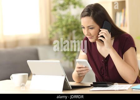 Imprenditore lavorando sulla linea con telefoni e tablet pc seduti a un tavolo nel soggiorno di casa con una finestra in background Foto Stock