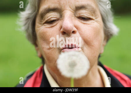 Donna anziana soffiando tarassaco in posizione di parcheggio Foto Stock