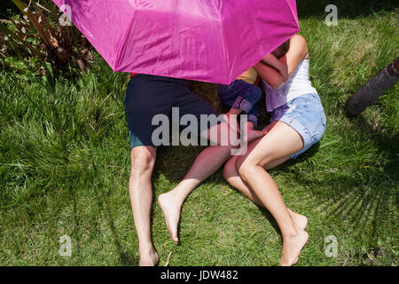 Genitori e figlio giacente su erba sotto ombrellone rosa Foto Stock