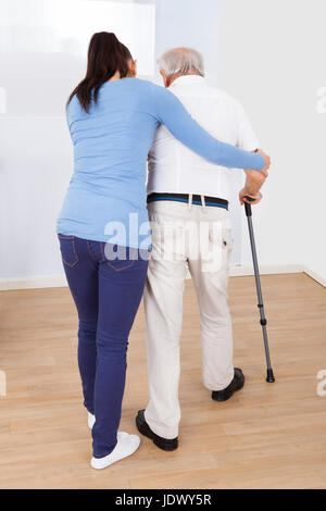 Lunghezza piena vista posteriore del caregiver assistenza disabili uomo senior a camminare con il bastone a casa di cura Foto Stock
