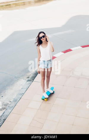 Slim giovane sportivo bella donna in bianco t-shirt, jeans brevi cortometraggi e alla moda di occhiali da sole blu ride shortboard penny skateboard. Scena urbana, città Foto Stock