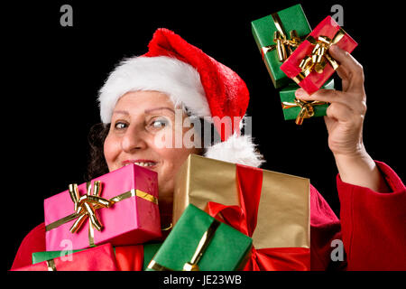 Allegra donna invecchiato con Babbo Natale cappuccio è offrendo tre piccoli regali di Natale in rilievo la sua mano sinistra. Più regali impacchettati in oro, verde e rosso sono tenuto vicino al petto. Isolato su nero. Foto Stock