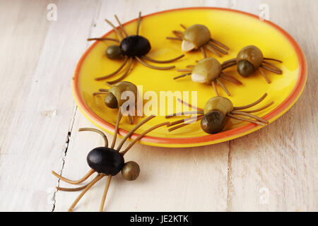 Creepy crawly Halloween spider snack per una celebrazione di partito fatta da olive nere e verdi italiana con gambe di spaghetti servita su una piastra di colore giallo su un bianco rustico tavolo in legno Foto Stock