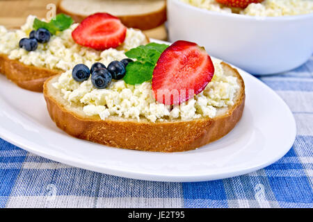 Fette di pane con crema di ricotta, menta, mirtilli e fragole, una ciotola di formaggio, pane su una scheda su uno sfondo di colore blu tovaglia a scacchi Foto Stock