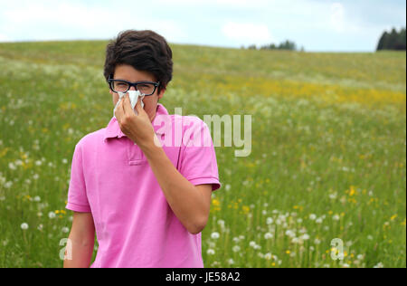 Allergia ragazzo con gli occhiali e t-shirt rosa soffia il naso con un fazzoletto nel mezzo di un campo Foto Stock