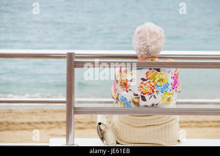 Donna anziana con i capelli bianchi e colorati top seduta sul banco di prova che si affaccia sul mare. Foto Stock
