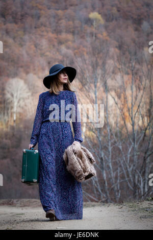 Piuttosto giovane donna con una valigia in pelle su un dirtroad in montagna, in stile vintage Foto Stock