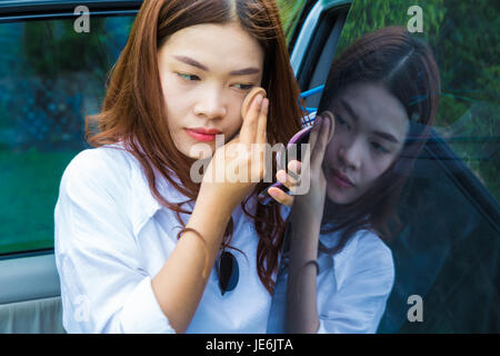 Attraente giovane donna asiatica, teen, mettendo make up sul suo volto, al di fuori della sua automobile con la sua riflessione sul vetro dell'auto Foto Stock