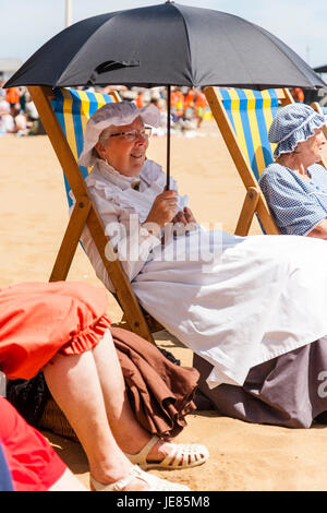 Senior donna, 60s, seduto in una sedia a sdraio, sulle sabbie a Broadstairs. Vestito in stile vittoriano cucina costume cameriera, azienda Ombrello nero contro il sole brillante. Sorridente. Vista laterale. Foto Stock