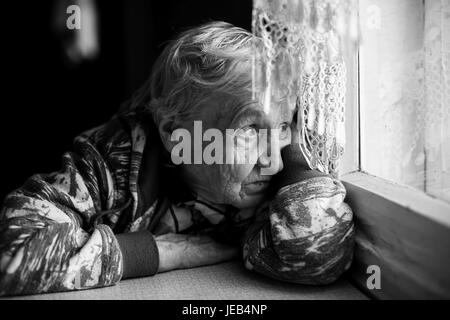 Una donna anziana wistfully guarda fuori dalla finestra. In bianco e nero photo. Foto Stock