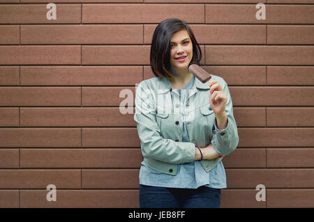 Bella sorridente giovane donna in un elegante giacca di pelle i gusti di gelato in glassa di cioccolato in prossimità di una superficie strutturata marrone a muro in mattoni. Foto Stock