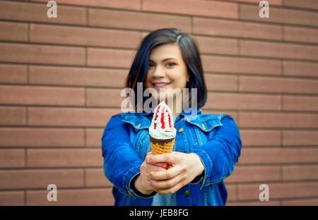 Bella caucasian brunette girl stretching le braccia in avanti con un gustoso gelato con confettura di frutta rossa in un jeans blu camicia vicino a una texture marrone Foto Stock