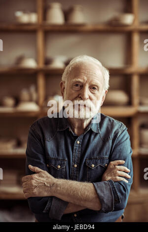 Ritratto di senior potter in piedi contro i ripiani con merci di ceramiche in officina Foto Stock