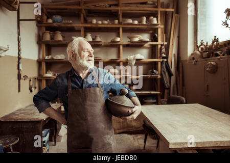 Vista frontale del fiero senior potter nel grembiule in piedi con la ciotola in ceramica in officina Foto Stock