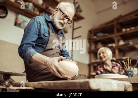 Senior potter nel grembiule e occhiali esaminando tazza ceramica con la donna lavora sullo sfondo Foto Stock