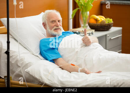 Sorridente senior paziente sdraiato nel letto di ospedale e mostrando il pollice in alto Foto Stock