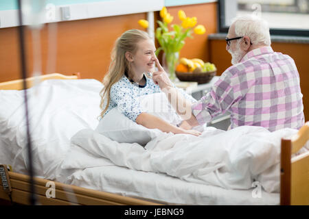 Senior uomo in occhiali visitando sorridente ragazza seduta nel letto di ospedale Foto Stock