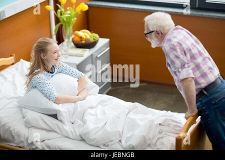 Senior uomo in occhiali visitando sorridente ragazza seduta nel letto di ospedale Foto Stock