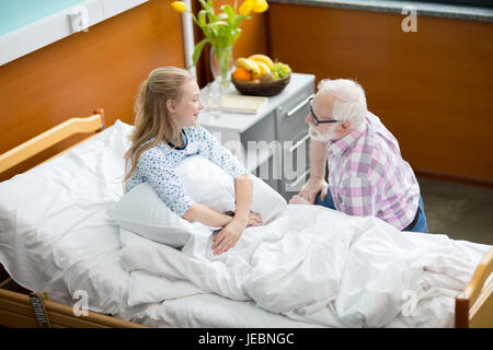 Senior uomo in occhiali visitando sorridente ragazza seduta nel letto di ospedale Foto Stock