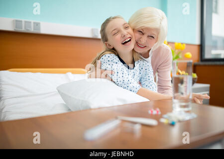 Nonna sorridente e nipote abbracciando comodamente seduti sul letto di ospedale Foto Stock