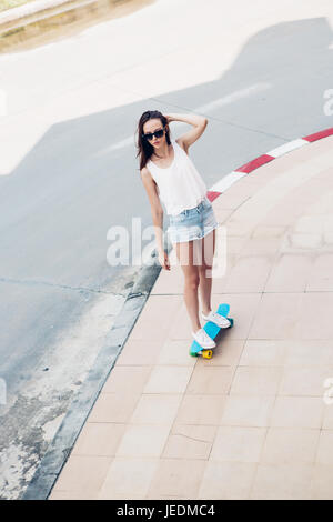 Slim giovane sportivo bella donna in bianco t-shirt, jeans brevi cortometraggi e alla moda di occhiali da sole blu ride shortboard penny skateboard. Scena urbana, città Foto Stock