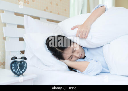 Giovane bella donna asiatica che giace nel letto che soffrono di sveglia sonora testa di copertura e le orecchie con cuscino rendendo faccia sgradevole. Early wake up, n. Foto Stock