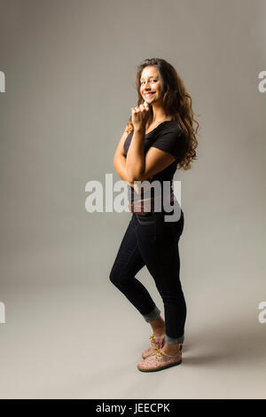 Giovani razza mista donna che indossa un nero di t-shirt e jeans si erge con fiducia con i capelli di soffiatura e mano sul mento mentre sorridente al viewer Foto Stock