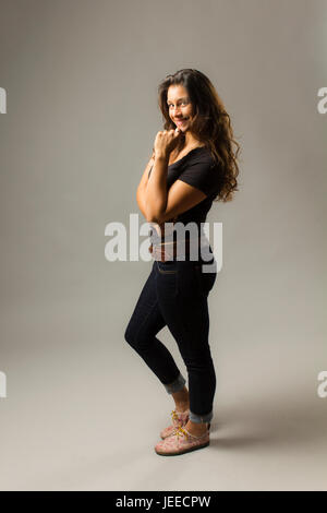Giovani razza mista donna che indossa un nero di t-shirt e jeans si erge con fiducia con i capelli di soffiatura e mano sul mento mentre sorridente al viewer Foto Stock