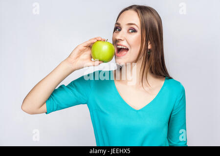 Giovane bella donna con le lentiggini e abito verde azienda apple e mangiare guardando la fotocamera. studio shot, isolato su sfondo grigio chiaro. Foto Stock