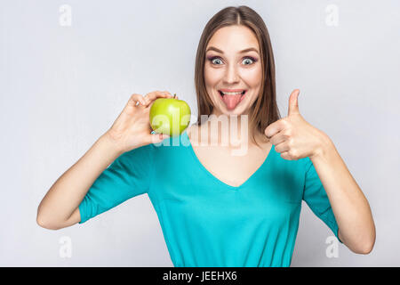 Giovane bella donna con le lentiggini e abito verde azienda apple con pollice in alto e la linguetta. studio shot, isolato su sfondo grigio chiaro. Foto Stock