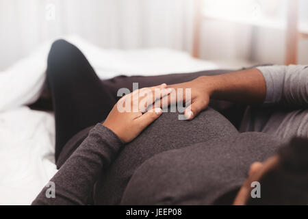 Close up di donne incinta giovane sdraiato sul letto di casa. Focus sulle mani di un uomo e di una donna su una gravidanza pancia. Foto Stock