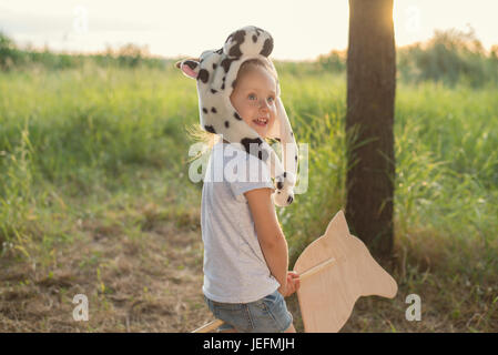 Kid in animali divertenti hat giocando con il giocattolo di legno sul cavallo tramonto Foto Stock